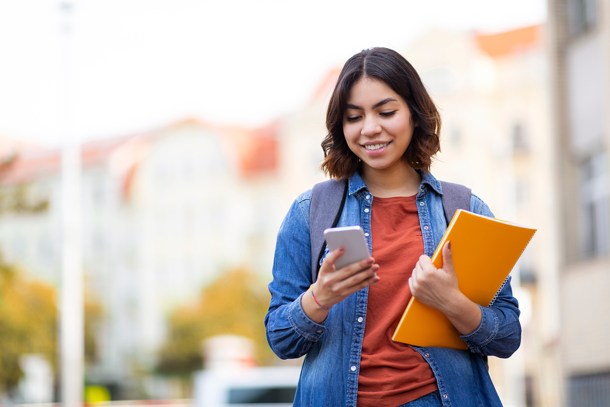 Jovem mulher de mochila, segurando um caderno e usando o celular.
