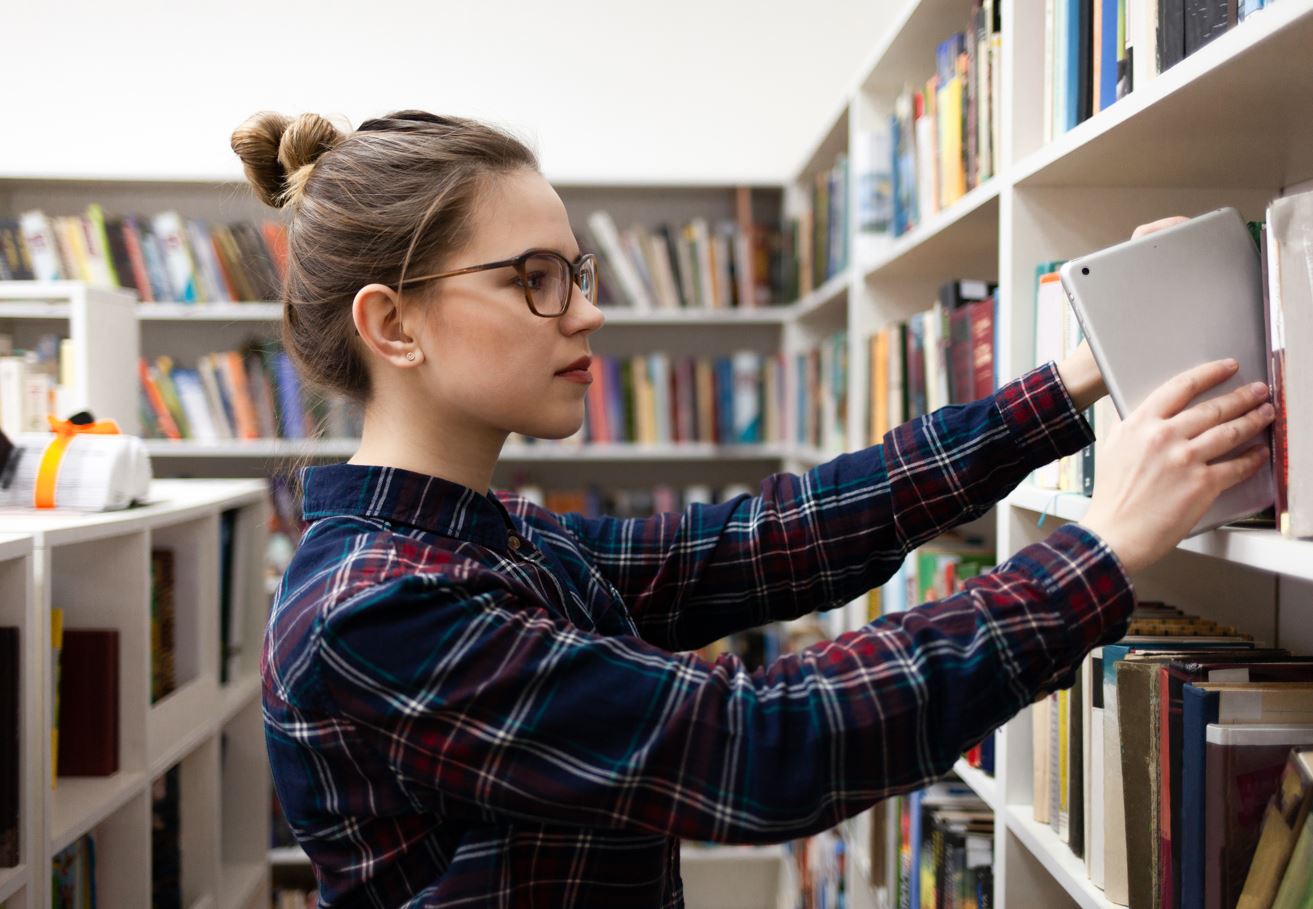 Jovem estudante coloca um tablet em uma prateleira com livros na biblioteca